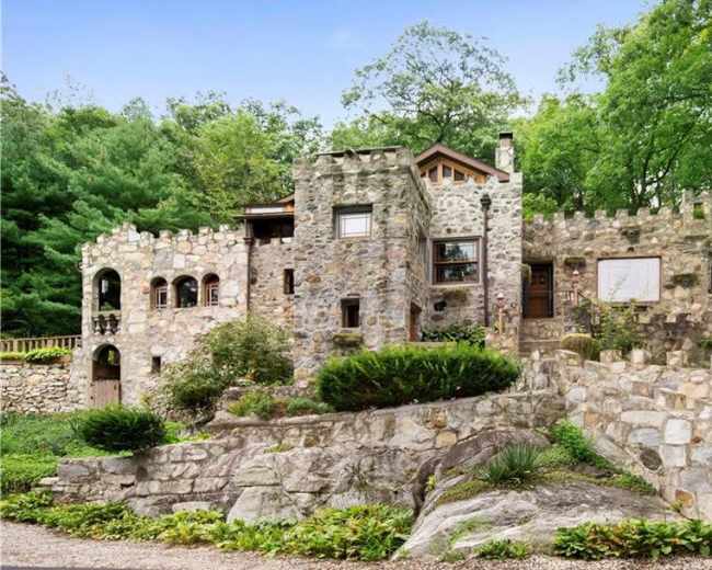 A stone house designed with castle-like features, surrounded by lush greenery and a landscaped pathway.