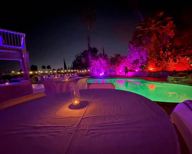 A table is set by a illuminated swimming pool surrounded by colorful lighting and palm trees at night.