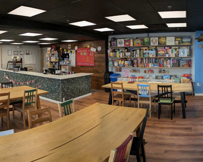 The image shows a brightly lit game café with multiple wooden tables, a counter displaying board games, and a bookshelf filled with various games along one wall.