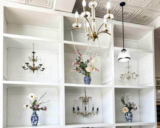 The image features a white shelving unit with various decorative items, including vases with flowers and chandeliers, arranged in a symmetrical pattern alongside colorful stools.