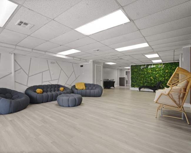 The image shows a spacious room with modern gray seating, light wooden flooring, and a green plant wall in one corner.