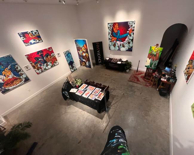 The image shows an art gallery featuring vibrant paintings on the walls and a display table with art prints in the center.