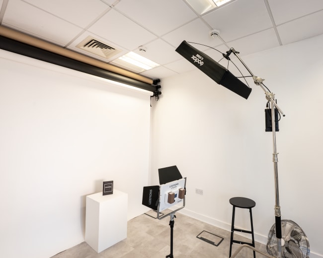 The image shows a minimalistic photography studio featuring a light setup, a stool, a pedestal for displays, and a fan against a white wall.