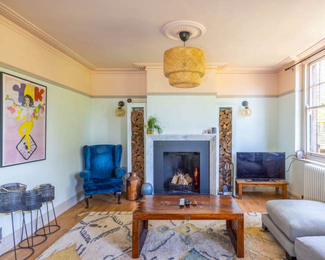 A living room features a blue armchair, a wooden coffee table, a television, a fireplace with stacked wood, and large windows allowing natural light.