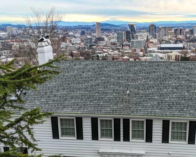 A white, two-story house with a gray shingle roof overlooks a city skyline featuring tall buildings and distant mountains.