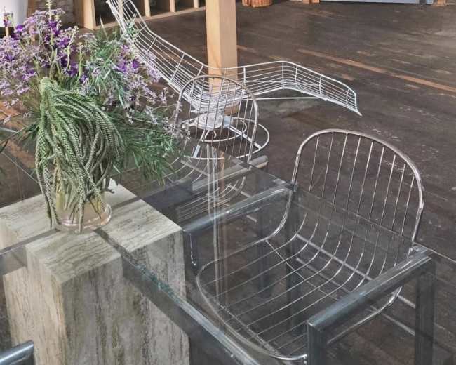 The image shows a spacious room with wooden floors, a brick wall, and two wire chairs positioned next to a glass table adorned with a vase of flowers.