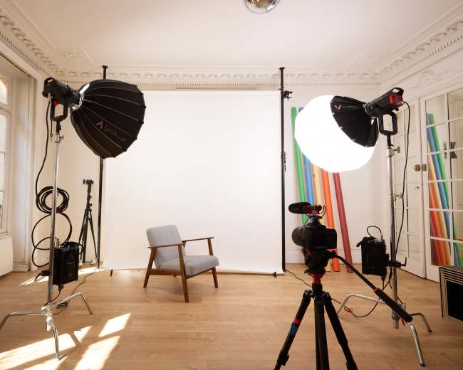 The image shows a photography studio with a gray armchair, camera equipment, and lighting setups positioned around a white backdrop.