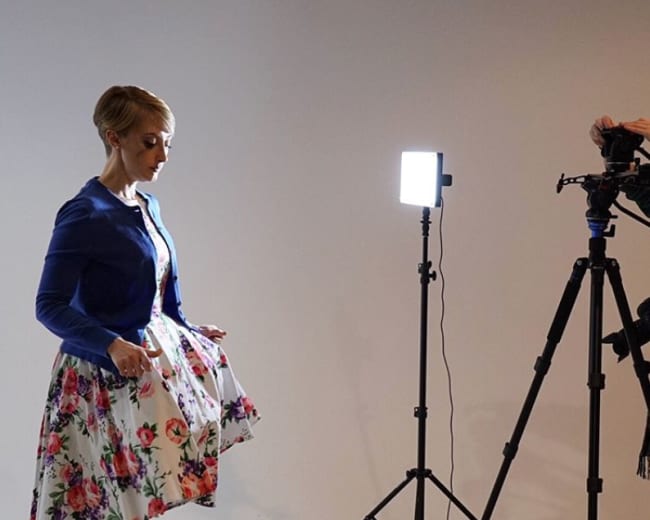 A woman in a floral dress adjusts her outfit in front of a camera set on a tripod, with a bright light illuminating the scene.