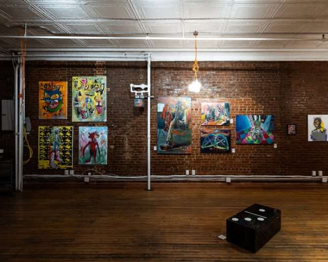The image shows an art gallery with exposed brick walls displaying colorful paintings and sculptures, illuminated by pendant lights.
