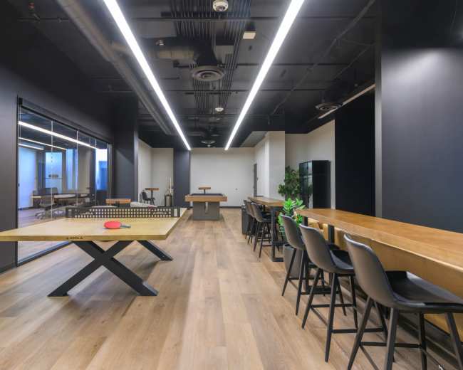 The image shows a modern office break room featuring a ping pong table, a bar-height table with seating, and plants, all set in a spacious, well-lit environment.