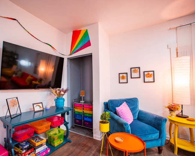 A brightly colored living room features a blue velvet chair, a television mounted on the wall, and shelves filled with vibrant storage bins.