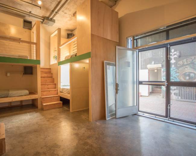 The image shows a modern interior space featuring two elevated sleeping areas, wooden steps leading to one of the beds, and a large glass door that opens to the outside.