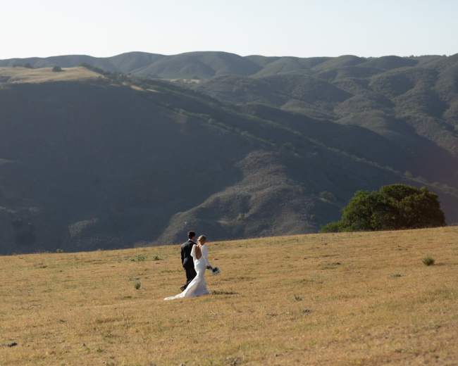 A couple walks together across a grassy field with rolling hills in the background.
