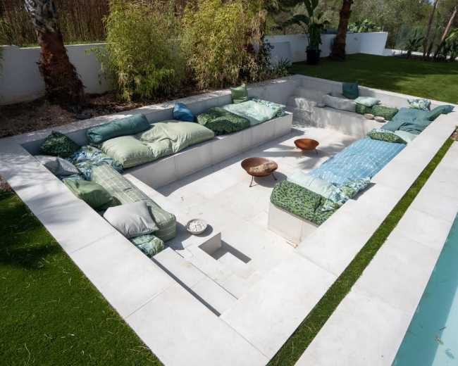 The image shows a modern outdoor seating area arranged in a sunken design, surrounded by lush greenery and adjacent to a swimming pool.