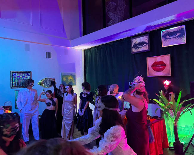 A group of elegantly dressed guests gathers in a brightly lit room adorned with vibrant colors and framed portraits, preparing for a themed event.