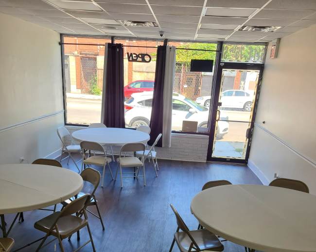 The image shows a vacant room with several round and rectangular tables set up, surrounded by chairs, and a large window letting in natural light.