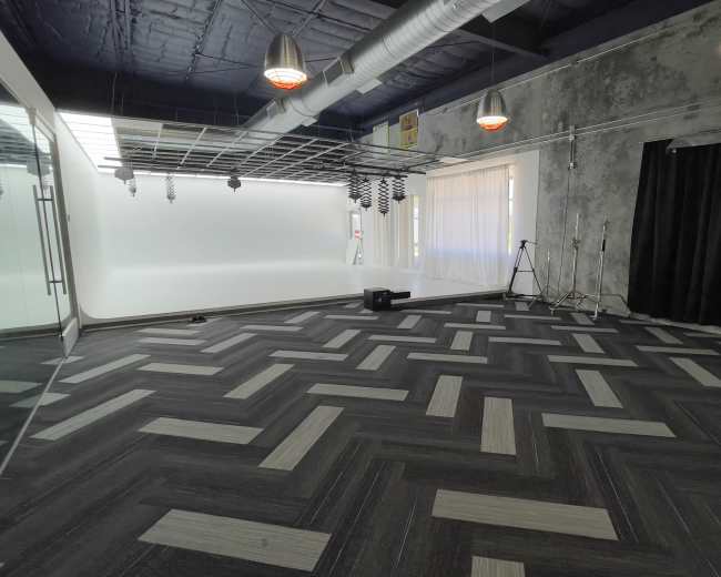 A spacious room with a patterned dark carpet, large windows with white curtains, and a backdrop area with studio lighting equipment.