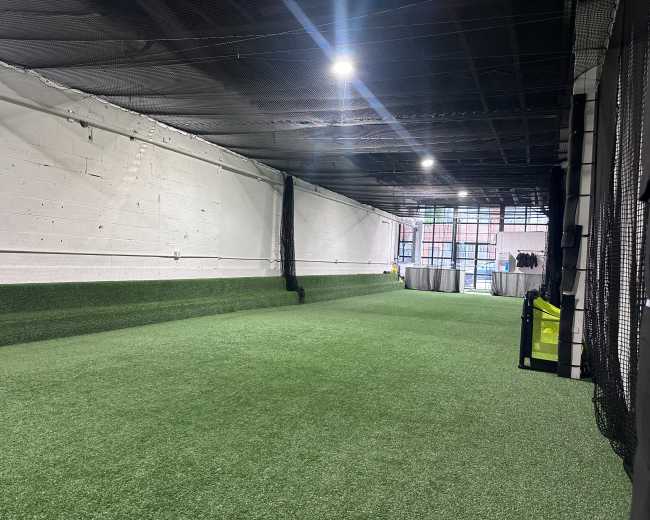 The image shows an indoor training space with artificial turf, high ceilings, and nets along the walls.