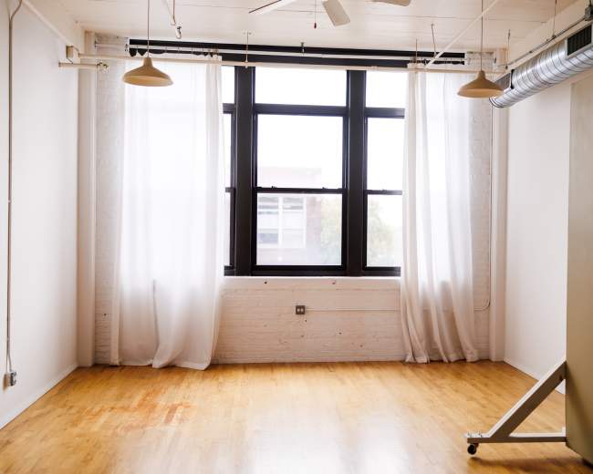 The image shows a minimalist interior space with large windows, sheer curtains, and a hardwood floor.