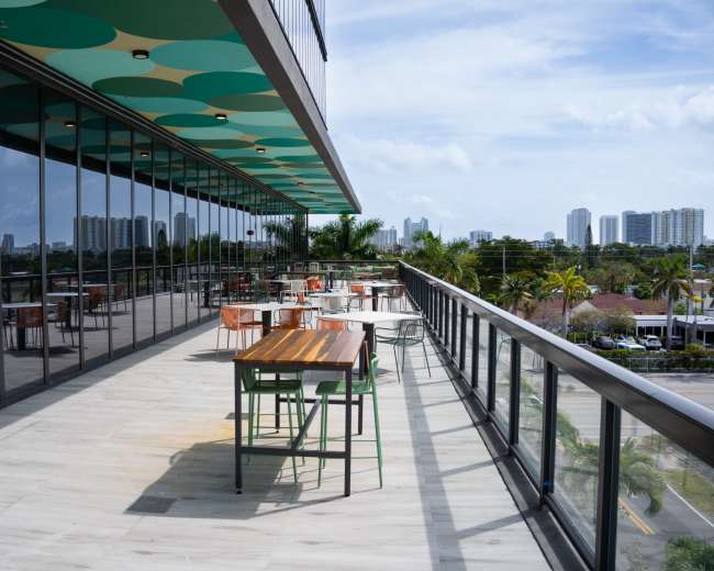 A rooftop terrace features several tables and chairs with a view of city buildings and a clear blue sky.