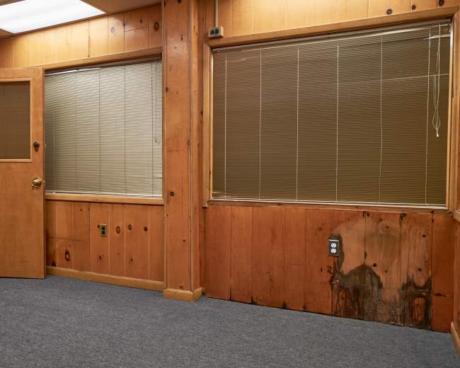 The image shows an interior space with wooden paneling, a door on the left, and two windows with blinds, featuring visible water damage on the wall beneath one of the windows.