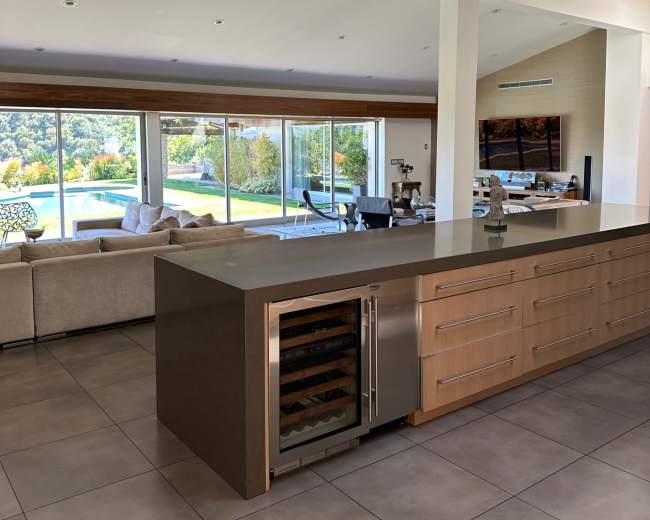 The image shows a modern kitchen with a sleek island and a view of a spacious living area that opens to an outdoor patio.