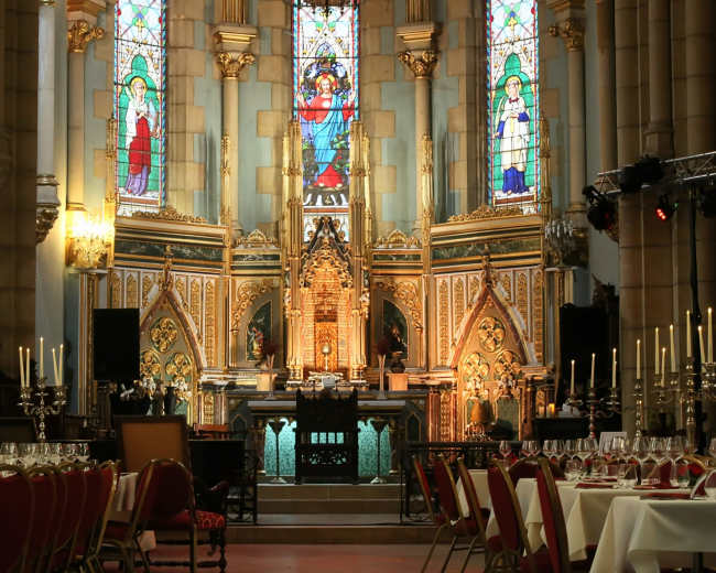 The interior of a cathedral features a dining setup with elegantly arranged tables and chairs in front of a beautifully ornate altar adorned with stained glass windows.