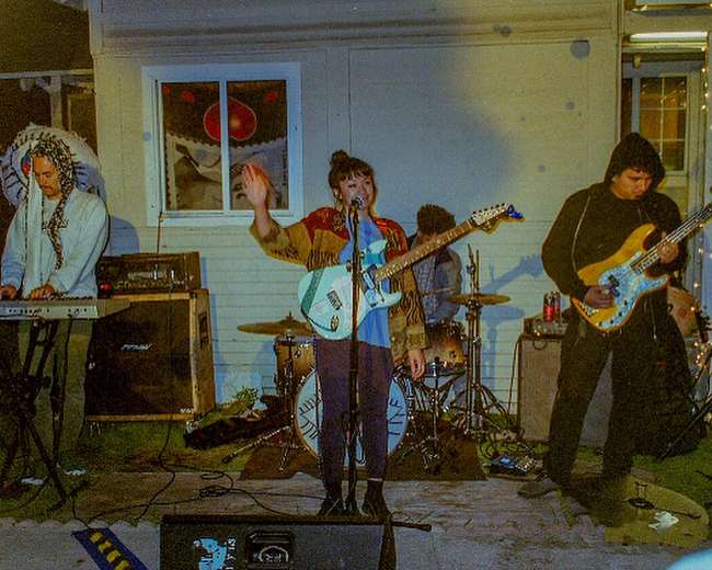 A band performs on an outdoor stage, with a female guitarist singing while the rest of the members play instruments in the background.