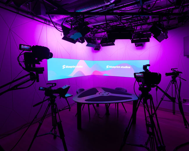 A studio space equipped with cameras and a large screen displaying the logo of "blueprint studios," illuminated by purple lighting.