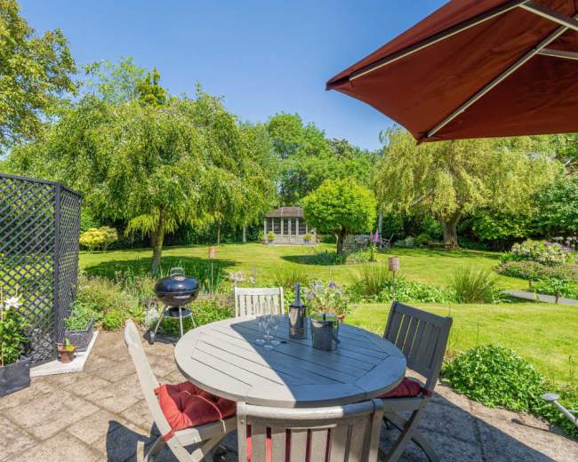 A patio table with chairs sits under an umbrella overlooking a landscaped garden with trees and a small shed in the distance.