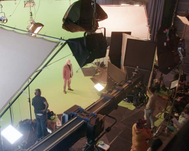 A film set features a green screen with various lights and equipment, while crew members prepare for a shoot.