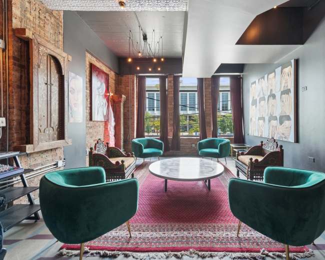 The image shows a stylish lounge area with modern green chairs, a round marble table, and artwork displayed on the walls, illuminated by natural light from large windows.