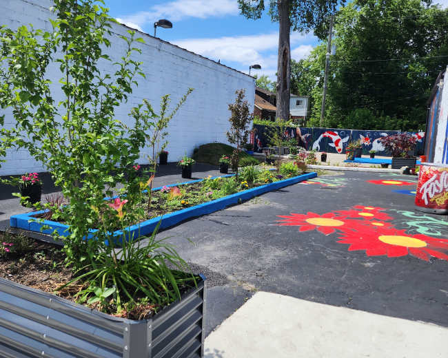 The image shows a vibrant community garden with flower beds and planters surrounded by a painted asphalt pathway and greenery under a clear blue sky.