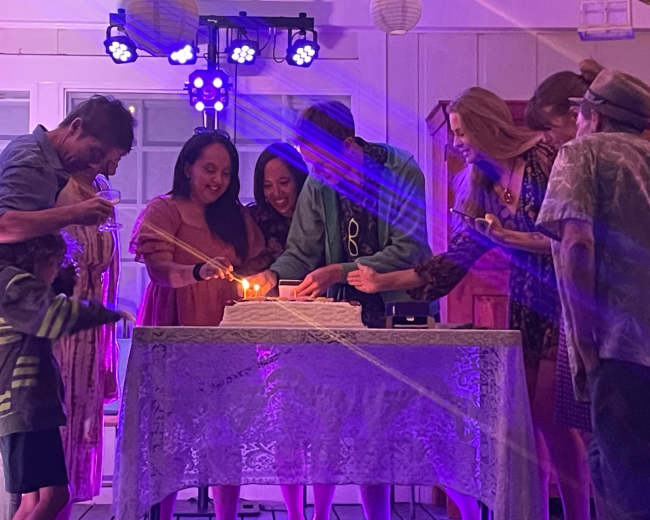 A group of people gathers around a cake, celebrating and lighting candles, under colorful lights in a decorated indoor space.