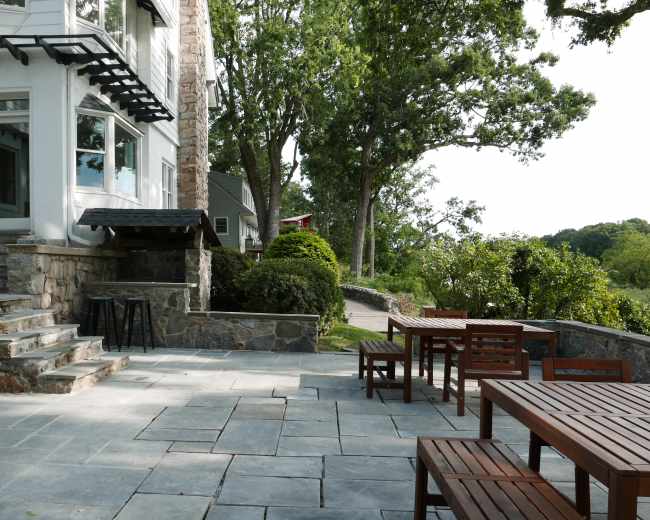 A stone patio with wooden tables and chairs is situated next to a house, surrounded by greenery and leading toward a pathway.