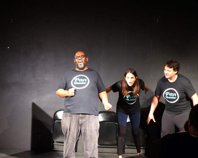 Three performers take the stage in black t-shirts with the "Pan Theater" logo, engaging in an energetic improv scene.