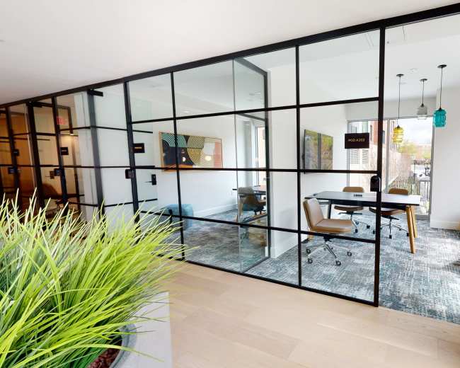 A modern office space is divided by glass walls, featuring a meeting area with a conference table and chairs, and accented by green plants outside.