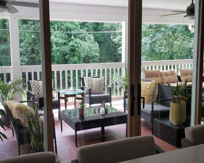 The image shows a covered porch with comfortable seating, a coffee table, and greenery visible through large sliding glass doors.