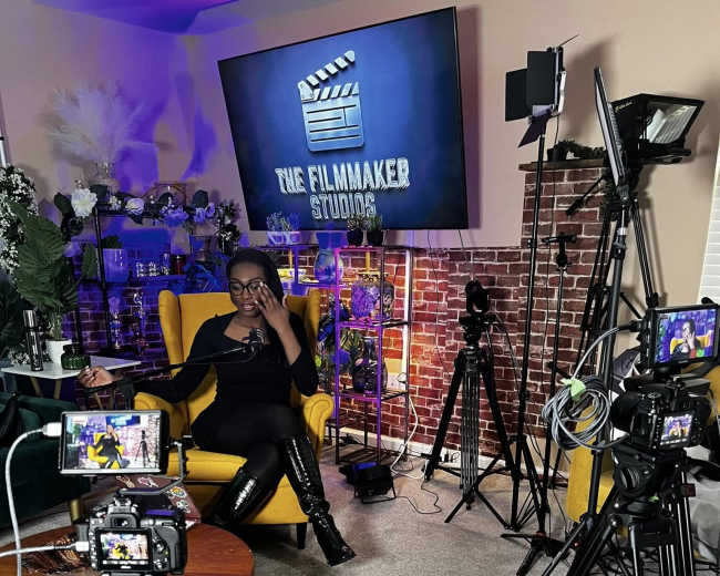 A woman sits in a yellow armchair in a studio setting, surrounded by multiple cameras and film equipment, with a large screen displaying “THE FILMMAKER STUDIO” in the background.