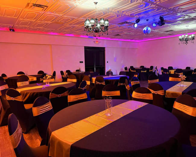 The image shows a banquet hall set up with round tables covered in black and gold tablecloths, surrounded by chairs adorned with gold sashes under colorful lighting.
