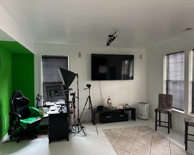 The room features a green accent wall, a desk with a computer and camera setup, a television mounted on the wall, two chairs, and a small area rug on a white floor.