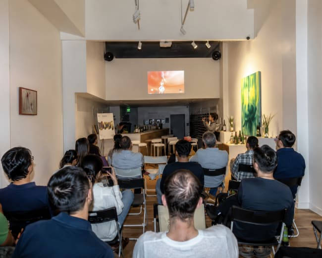 A group of people sits in chairs facing a screen in a modern, bright room with a bar and art on the walls.