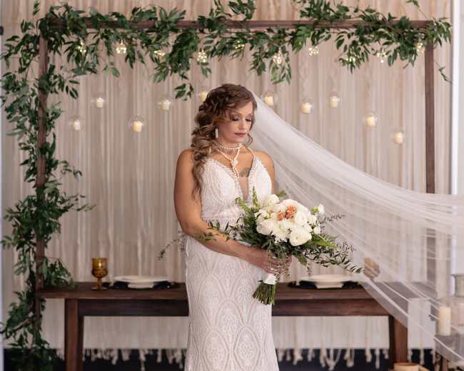 A bride in a lace wedding dress holds a bouquet of flowers while standing in front of a decorated altar with greenery and lights.
