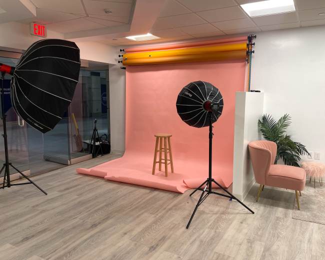 The image shows a photography studio setup featuring a pink backdrop, a wooden stool, two softbox lights, and a pink chair with a decorative plant nearby.
