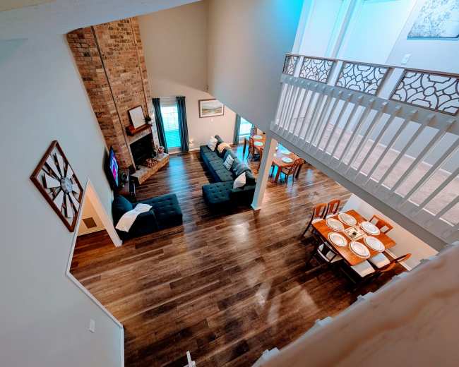 The image shows a spacious living area featuring a brick fireplace, two long sofas, and a dining table set for several people.