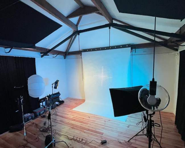 A photography studio features a brightly lit backdrop, camera equipment, and various lighting setups on polished wooden floors.