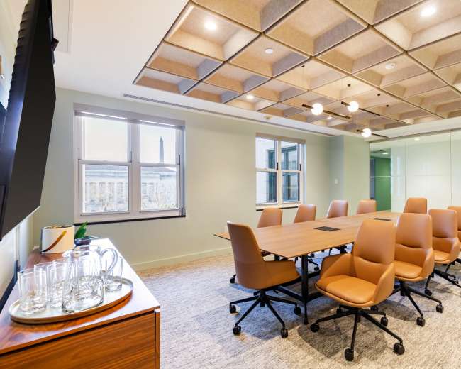 The image shows a modern conference room featuring a large wooden table surrounded by brown ergonomic chairs, with a glass wall and a plant in the corner.