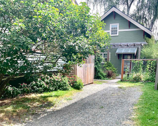 A gravel pathway leads through greenery toward a green house with a porch.
