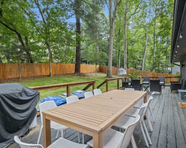 A wooden deck features a large table surrounded by white chairs, with a barbecue grill and a view of a tree-lined yard behind.
