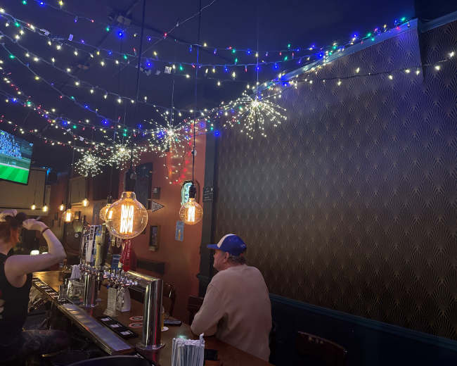 The image shows a bar with colorful string lights overhead, featuring a lone patron sitting at the counter.